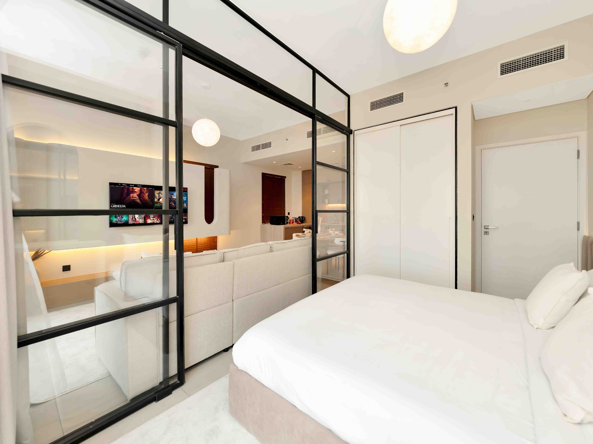 Bedroom view through full-height steel-framed glass partition to living room and kitchen beyond